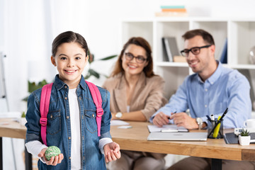 selective focus of cheerful kid with backpack holding ball near happy parents in office