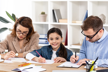 cute child drawing near parents in glasses working in office