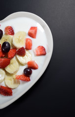 Half Heart-shaped cream plate filled with yogurt with banana slices, strawberries cut into pieces and grape berries on a black background. "Indulgence for breakfast" concept.