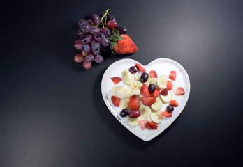 Heart-shaped cream plate filled with yogurt with banana slices, strawberries cut into pieces and grape berries on a black background. 