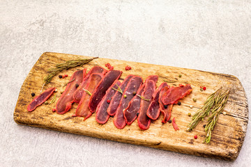 Basturma, dried tenderloin of beef meat, jerky, thinly sliced. Dry rosemary, pepper mix on vintage wooden board. Delicious food on a stone background.