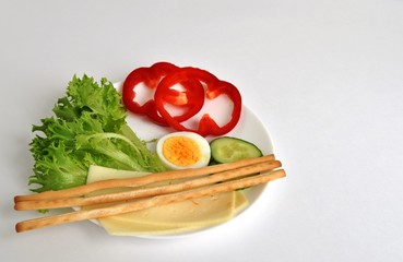 hard cheese, cucumber, egg, lettuce, pepper, bread sticks on a white plate and flower background