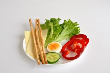 hard cheese, cucumber, egg, lettuce, pepper, bread sticks on a white plate and flower background