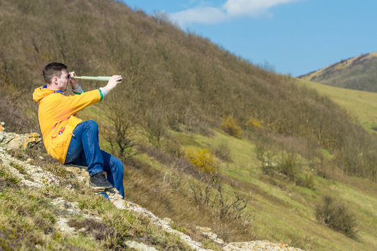 A young man in a yellow jacket, blue jeans sits on a rock and looks into a spyglass. In the background are mountains and sky. Mountain landscape.