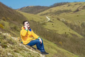 Naklejka na ściany i meble A young man in a yellow jacket, blue jeans and glasses sits in the mountains and enjoys the scenery and talking on the phone, laughing. In the background are hills and sky. Mountain landscape.