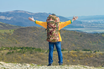A young man in a yellow jacket, blue jeans and a hat with a large travel backpack is standing on the mountain, with his back to the frame and holding up his hands, a sign of freedom. 