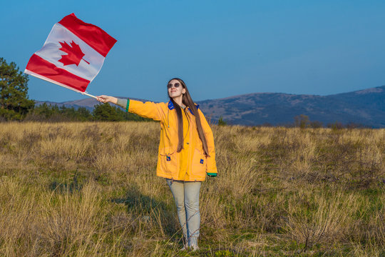 A young girl in a yellow jacket, gray pants and glasses holds in her hand the flag of Canada. In the background are hills and sky. The flag of Canada is developing in the wind. - Powered by Adobe