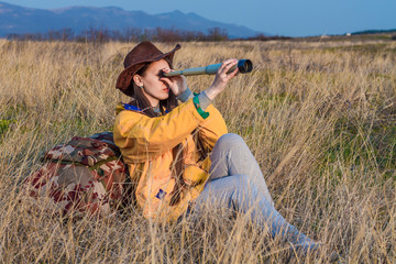 A young girl with long dark hair in a yellow jacket and a leather cowboy hat sits in the grass, leaning on a tourist backpack and looking through a telescope. Background mountains, sky.