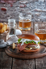  burger with bacon and cutlet with cheese, tomato, greens. served with fries and beer on a wooden table