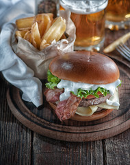  burger with bacon and cutlet with cheese, tomato, greens. served with fries and beer on a wooden table