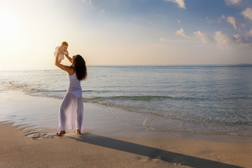 Happy mother holds her baby girl up in the air on a beach during sunset time