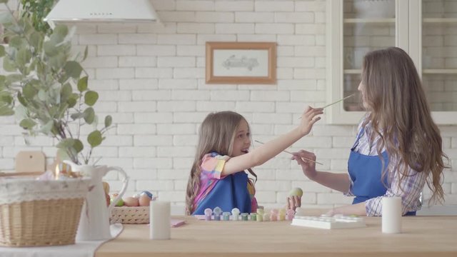 Little girl painting eggs with her mother using a small brush. The kid trying to touch woman' s nose with a brush. Preparation for Easter holiday. Mother and daughter have fun together in the kitchen