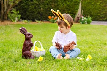 Cute little baby boy with bunny ears eating chocolate Easter bunnies sitting on green grass outside...