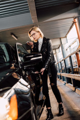 Fototapeta premium blonde attractive girl looking at smartphone while standing near car