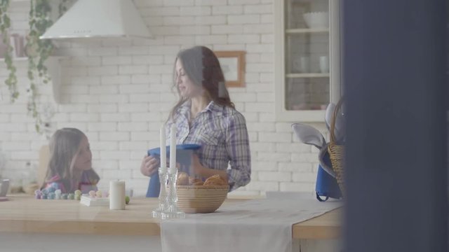 Woman taking blue apron from the basket and giving it to little girl in the kitchen. Preparation for Easter holiday. Acrylic paint bottles on the table, ladies going to paint eggs. Camera moves left