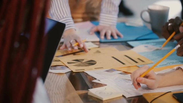 Close-up multiethnic business partners collaborate, brainstorm over large office table with printed marketing charts.