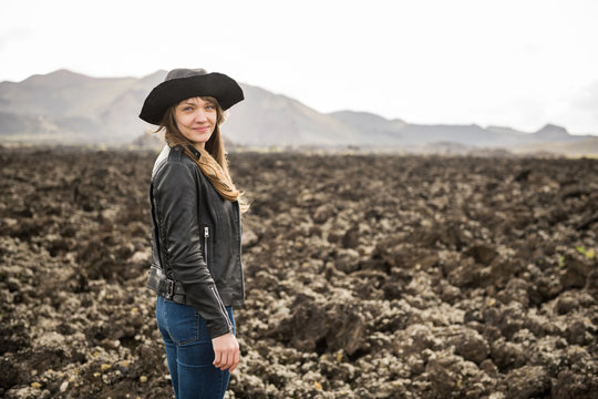 Side view of attractive female in casual outfit and hat smiling and looking at camera while standing on rocky ground in majestic countryside