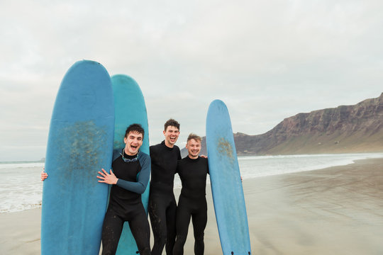 Three young males in black swimwear laughing and looking at camera while holding blue surfboards and standing on shore near sea