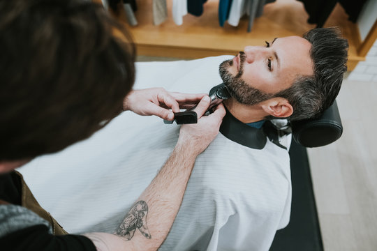 barber with comb and trimmer cutting beard of male sitting in barbershop on blurred background