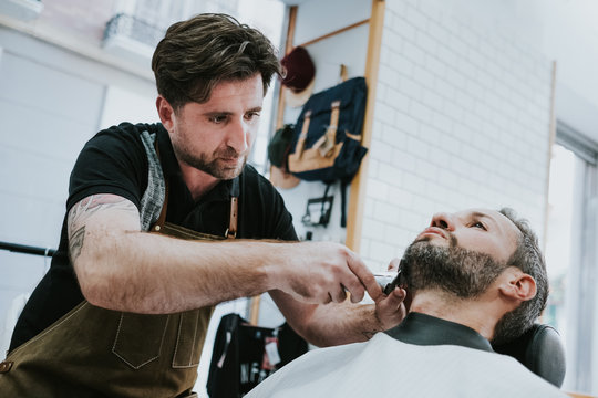 barber with comb and trimmer cutting beard of male sitting in barbershop on blurred background