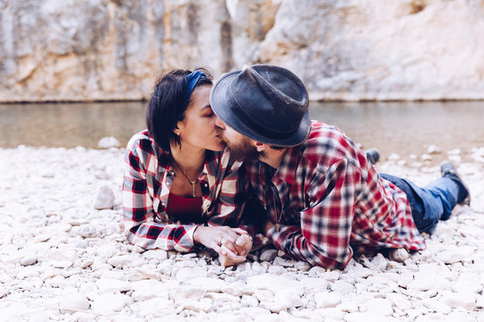 Side view of young couple lying and kissing on rock coast of mountain river near cliff
