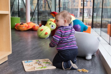 Horizontal shot of cute fair baby girl seen in profile kneeling in playroom full of giant soft toys and colourful poufs playing with wooden puzzle - Powered by Adobe