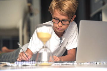 Handsome blond kid eyeglasses doing homework with computer keeping eye time available hourglass. healthy child searching information internet; Virtual classroom; education, home-schooling concept;