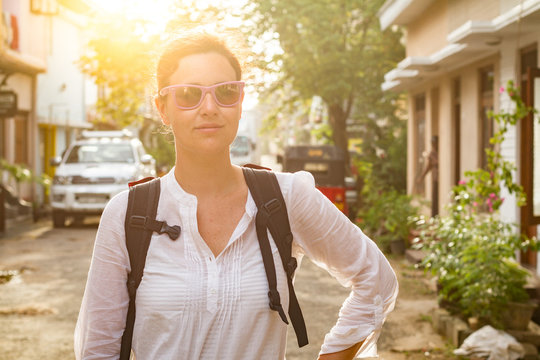 Attractive woman with backpack standing on road near lovely buildings and green plants in small town in Sri Lanka