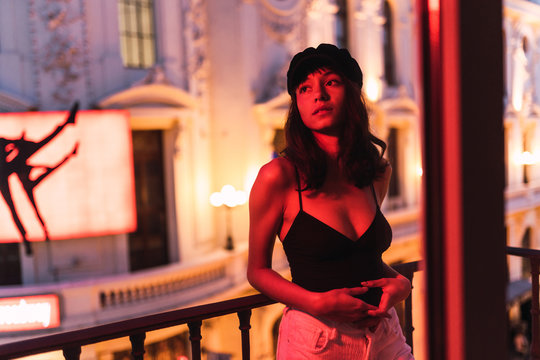 Young slim woman in cap standing near balcony in room in redness at night