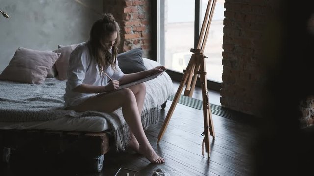 girl artist paints a picture of sitting on the bed in the shirt. painter works in a home studio in the morning.