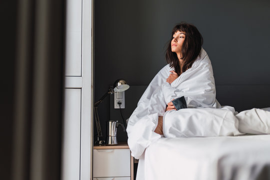 Side view of young dozy woman with mug of drink in duvet sitting on bed in bedroom
