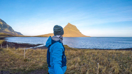 A young man wearing blue jacket and a beanie walks around famous Kirkjufell mountain in Iceland. Sea surrounding the hat like mountain. Beautiful and clear weather. Man taking a selfie