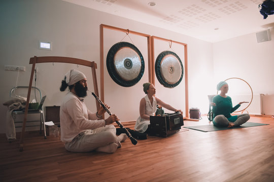 Woman sitting in lotus pose near musicians playing on ethnic instruments near gongs in room