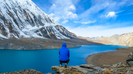 Young man squatting at the rock in front of Tilicho Lake. Blue and calm surface of the lake, mountains covered in the shadow, sunlight in the back. Overcast. Harsh landscape. Thankfulness, freedom.