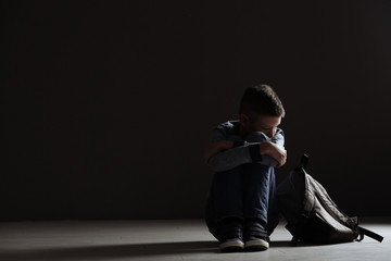 Upset boy with backpack sitting in dark room. Space for text