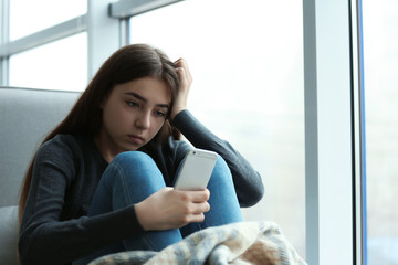 Upset teenage girl with smartphone sitting at window indoors. Space for text - obrazy, fototapety, plakaty