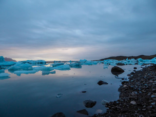 Beautiful glacier lagoon, with pebbly shore. Thousands of icebergs drifting lazily towards the sea, shining in many shades of blue. Soft sunset in the back. Thick clouds above the lagoon.
