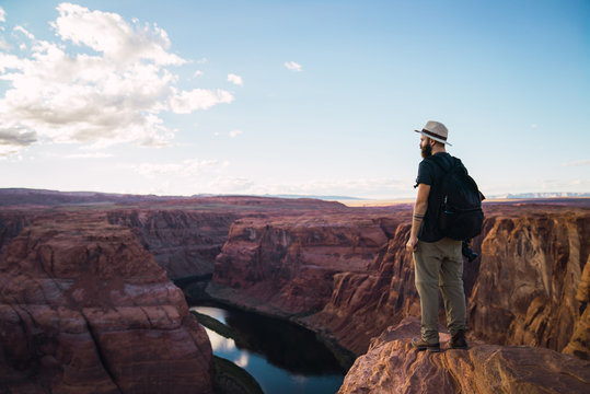 Side view of bearded guy with backpack holding photo camera to take pictures of beautiful canyon and calm river on sunny day on West Coast of USA