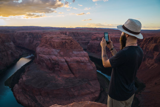 Handsome bearded man in hat taking picture while standing against magnificent canyon and river during sunset on West Coast of USA
