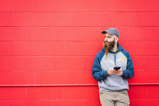 Attractive guy with braided beard browsing smartphone while leaning red wall on city street