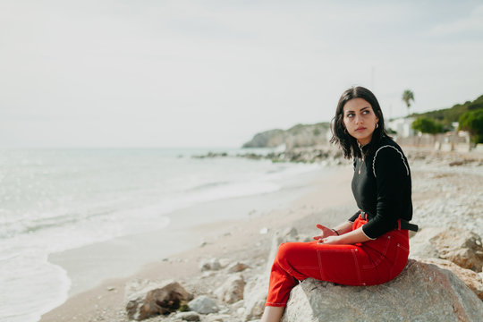 stylish woman looking away in calm sea while leaning on stones on sunny day