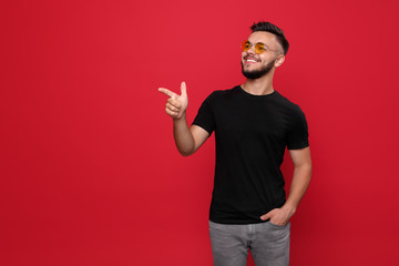 Bright bearded man in yellow sunglasses and black t-shirt pointing away laughing on red background.