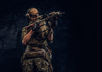 The elite unit, special forces soldier in camouflage uniform holding an assault rifle with a laser sight and aims at the target. Studio photo against a dark wall
