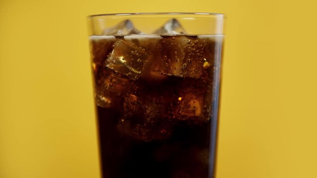 Glass full of cola fizzy drink with ice cubes. Cold coke sparkling soda on yellow background, moving bubbles slow motion