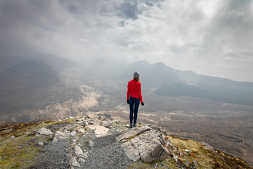 une jeune femme en rouge est au sommet d'une montagne face à un panorama sous les nuages