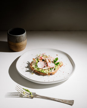 Trendy avocado toast topped with smoked trout fish, arugula, radish, sea salt, and black pepper. Served with coffee in a artisan hand made ceramic mug.