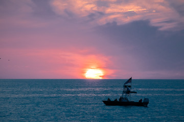A boat drifts in the ocean in front of a glowing setting sun amid the blue and purple clouds of evening, as seen from a beach on the Gulf of Mexico near Englewood, Florida, USA, in early spring