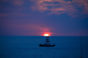 A boat on the blue ocean water in front of a glowing setting sun amid the blue and purple clouds of evening, as seen from a beach on the Gulf of Mexico near Englewood, Florida, USA, in early spring