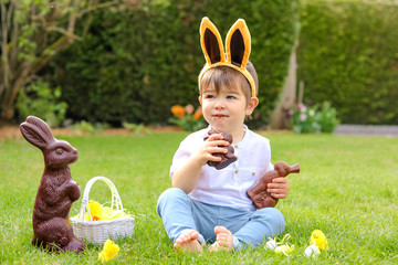 Cute little baby boy with bunny ears eating chocolate Easter bunnies sitting on green grass outside in the spring garden with basket of easter eggs and big chocolate rabbit. Happy sweet childhood