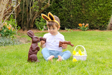 Cute little baby boy with bunny ears on head holding  chocolate Easter bunnies sitting on green grass outside in the spring garden with basket of easter eggs looking at big chocolate rabbit. 